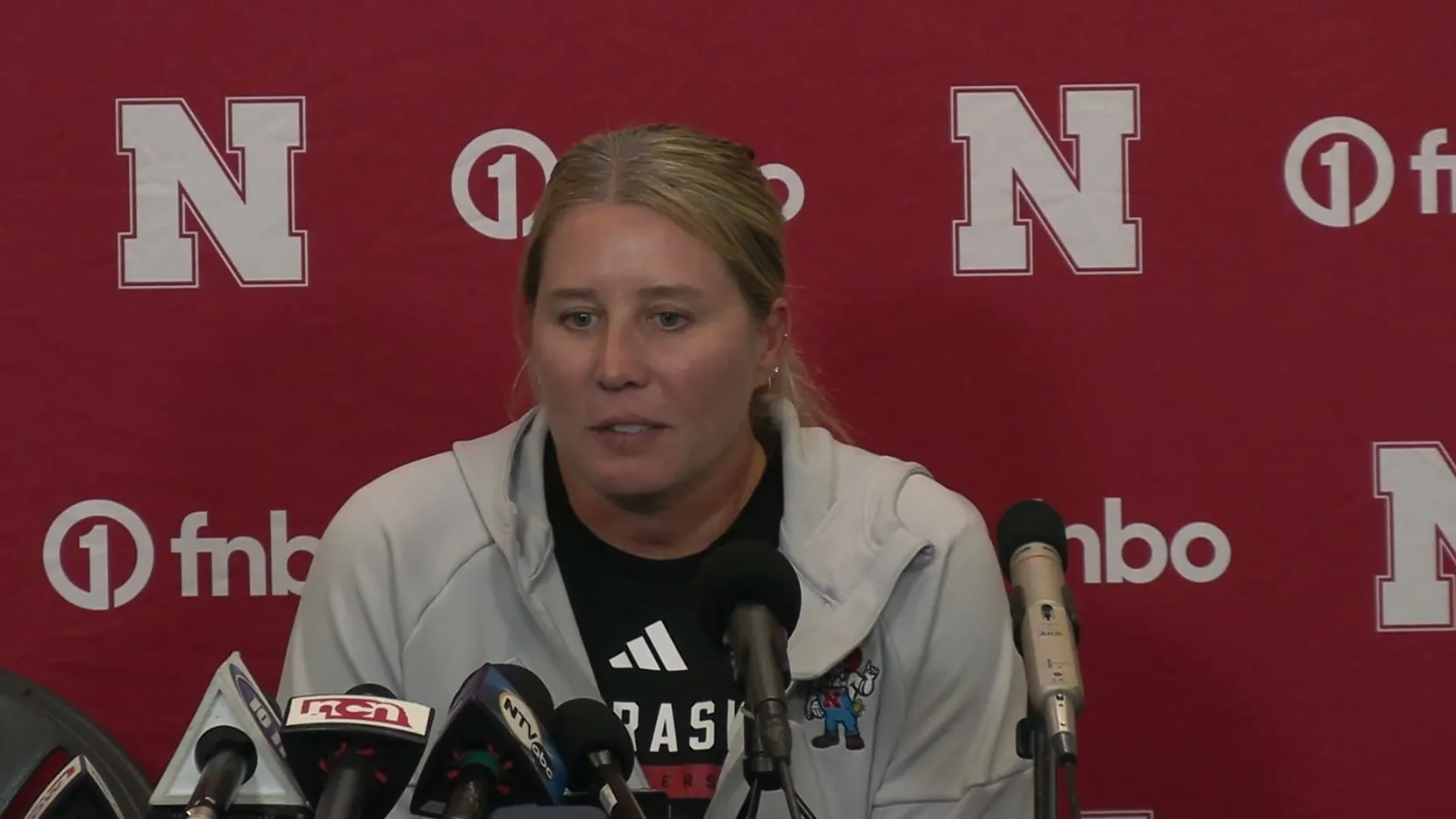 Dani Busboom Kelly speaking at the Nebraska press conference lectern behind a row of microphones.