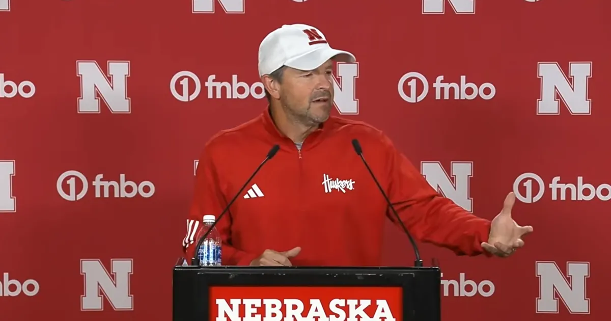 Glenn Thomas speaking at a podium marked Nebraska, wearing a red Huskers quarter-zip and a white cap, in front of a red backdrop.
