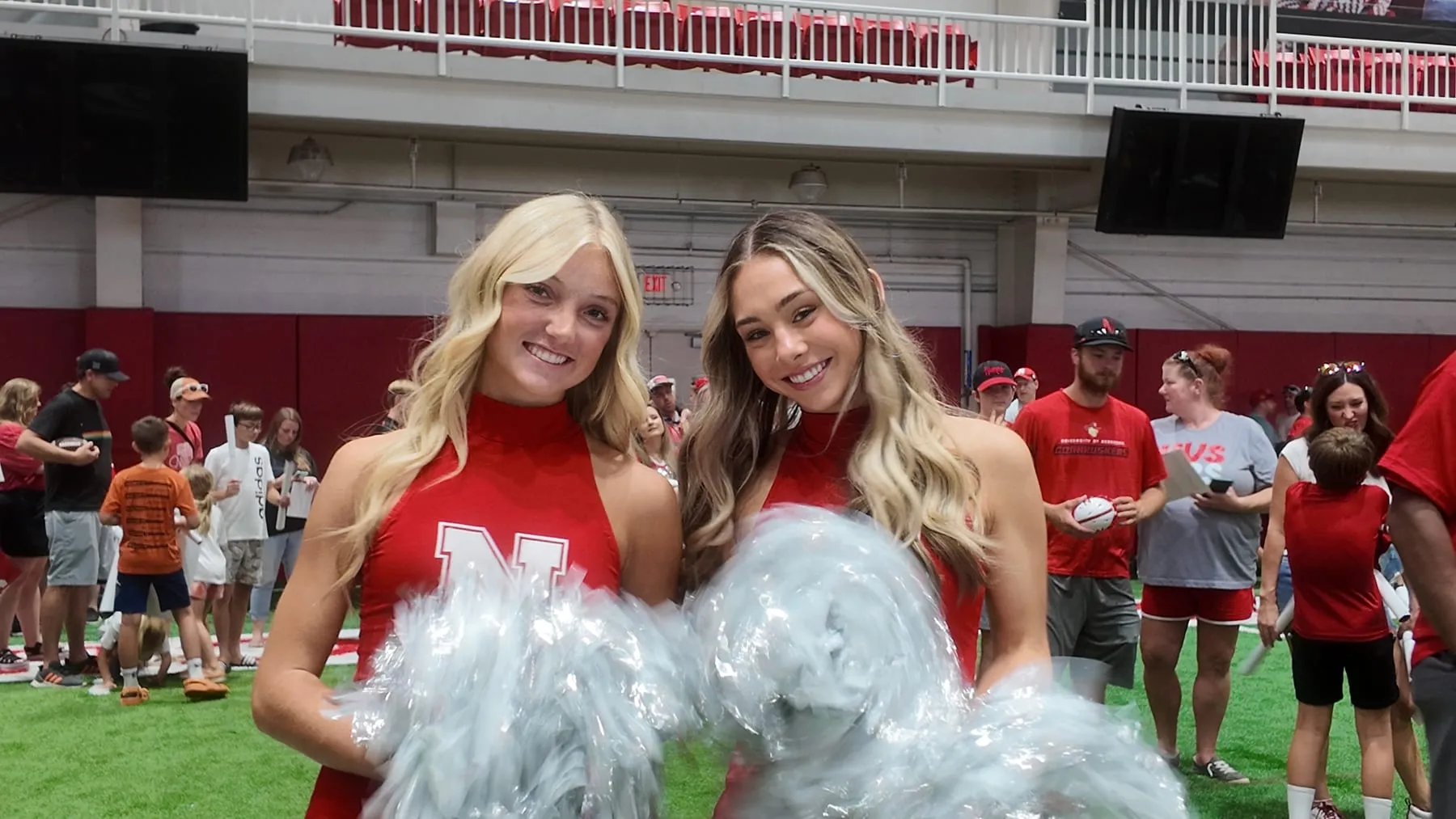 Two Nebraska cheerleaders in red uniforms hold silver pom-poms and smile at the camera while families and fans fill the indoor practice field behind them.