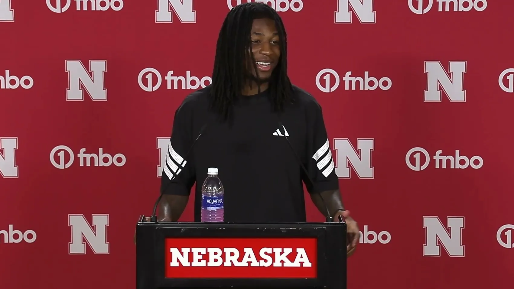 Jacory Barney Jr. speaking at the Nebraska press conference lectern in front of a red backdrop, mid-answer and smiling.