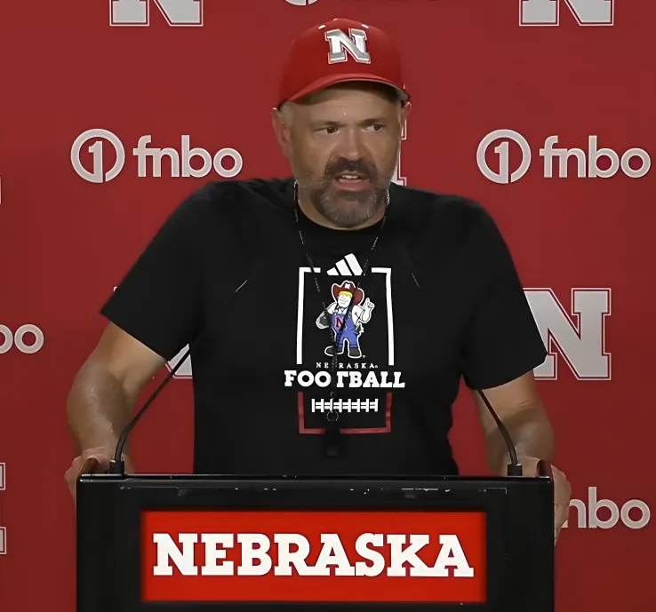 Matt Rhule speaking from a podium marked Nebraska, in a red Nebraska cap and a black Nebraska Football T-shirt, against a red backdrop.