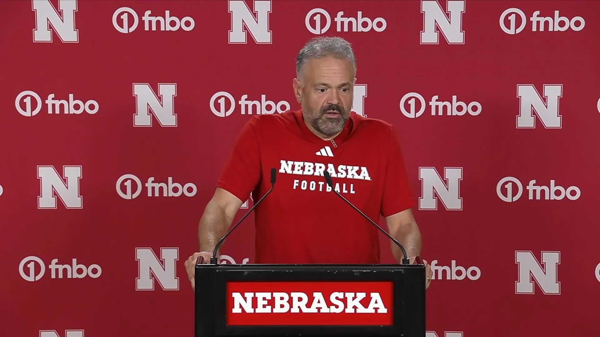 Nebraska head coach Matt Rhule in a red Nebraska Football shirt speaks at a podium in front of a red Nebraska and FNBO backdrop.