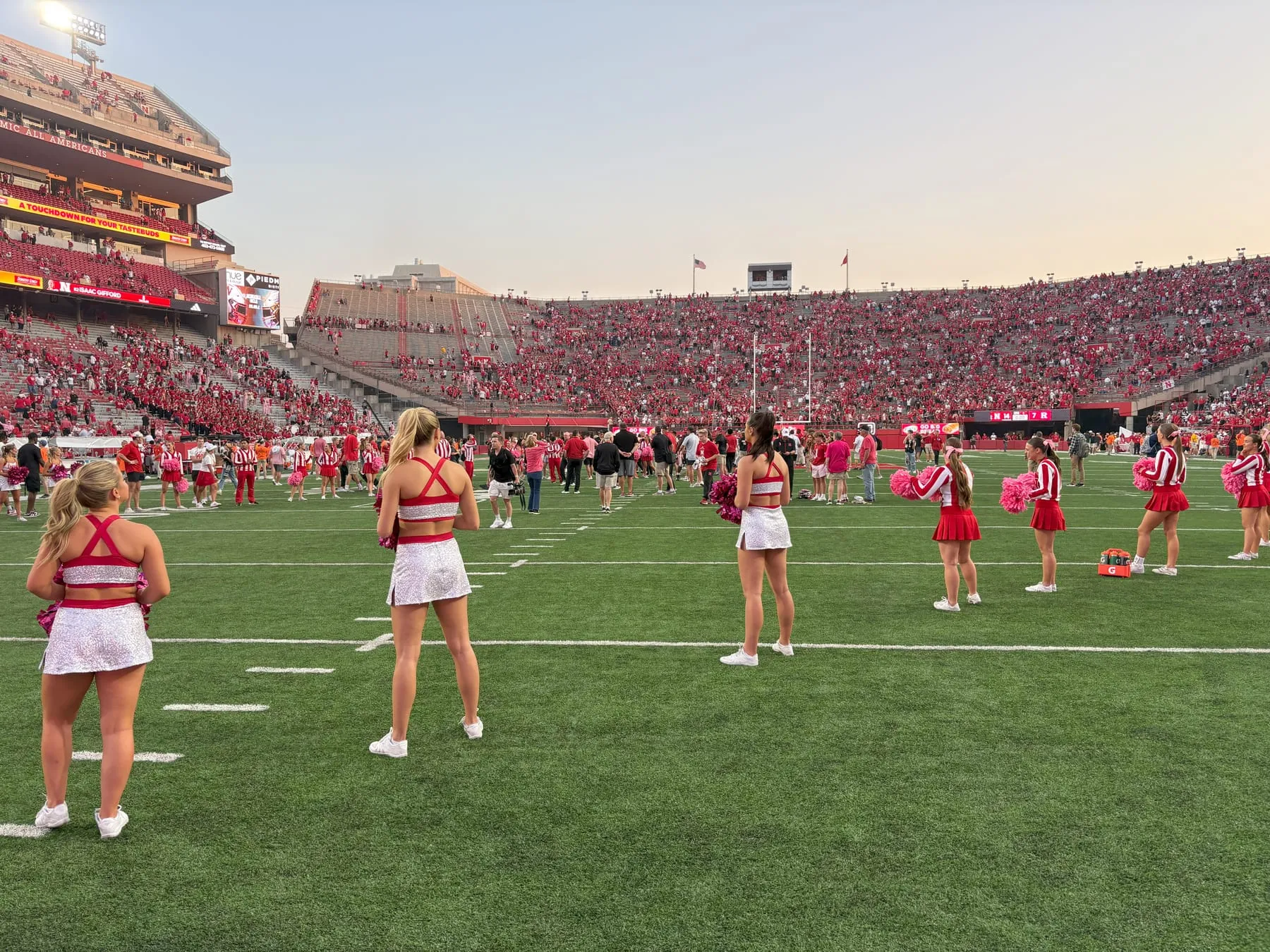 Nebraska cheerleaders stand along the sideline at dusk with Memorial Stadium full behind them.