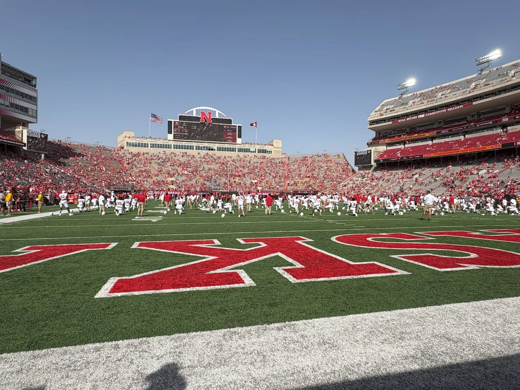 Memorial Stadium seen from field level before kickoff, both teams warming up across the HUSKERS end zone lettering with the stands filled in red.