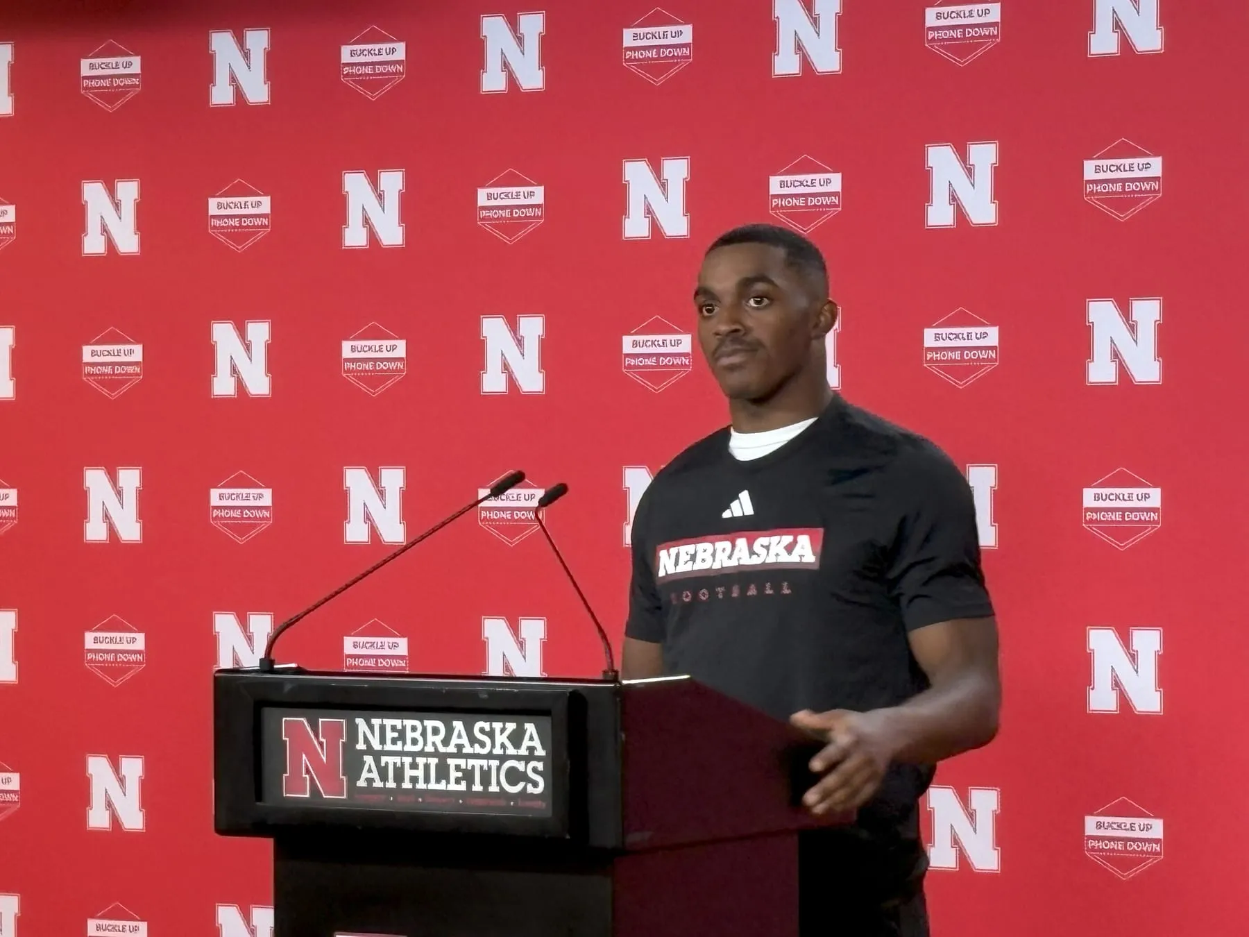 A Nebraska player speaks at a lectern in front of a red Nebraska Athletics backdrop.