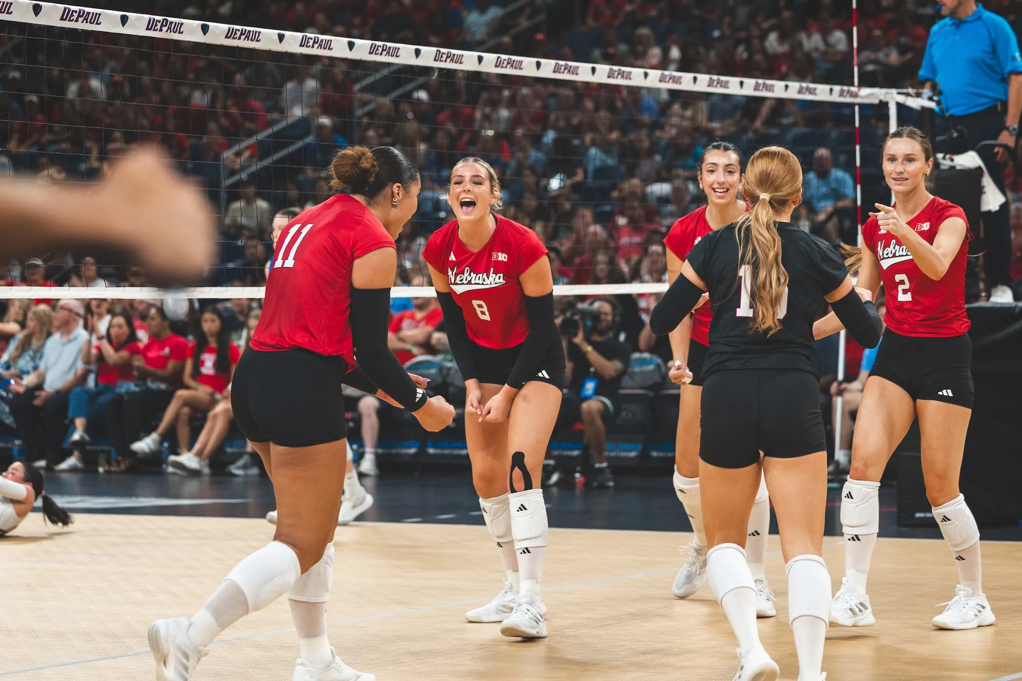 Nebraska players celebrate a point at the net against DePaul, with a packed Wintrust Arena crowd behind them.