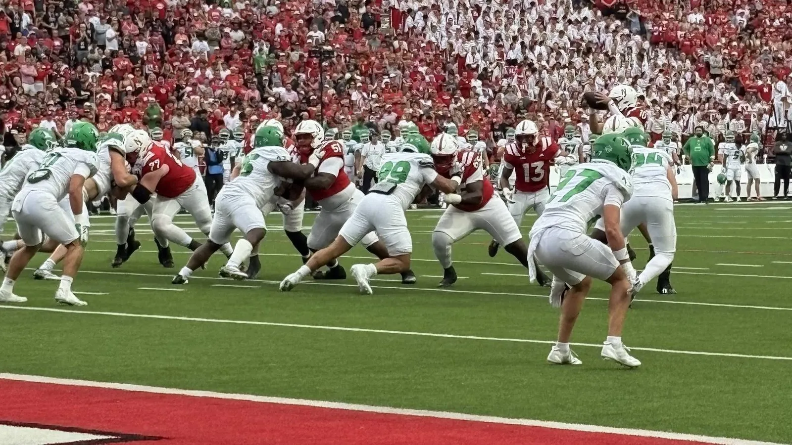 Nebraska's offensive line fires off the ball against North Dakota at Memorial Stadium, the quarterback holding the ball behind them and a full crowd in the background.