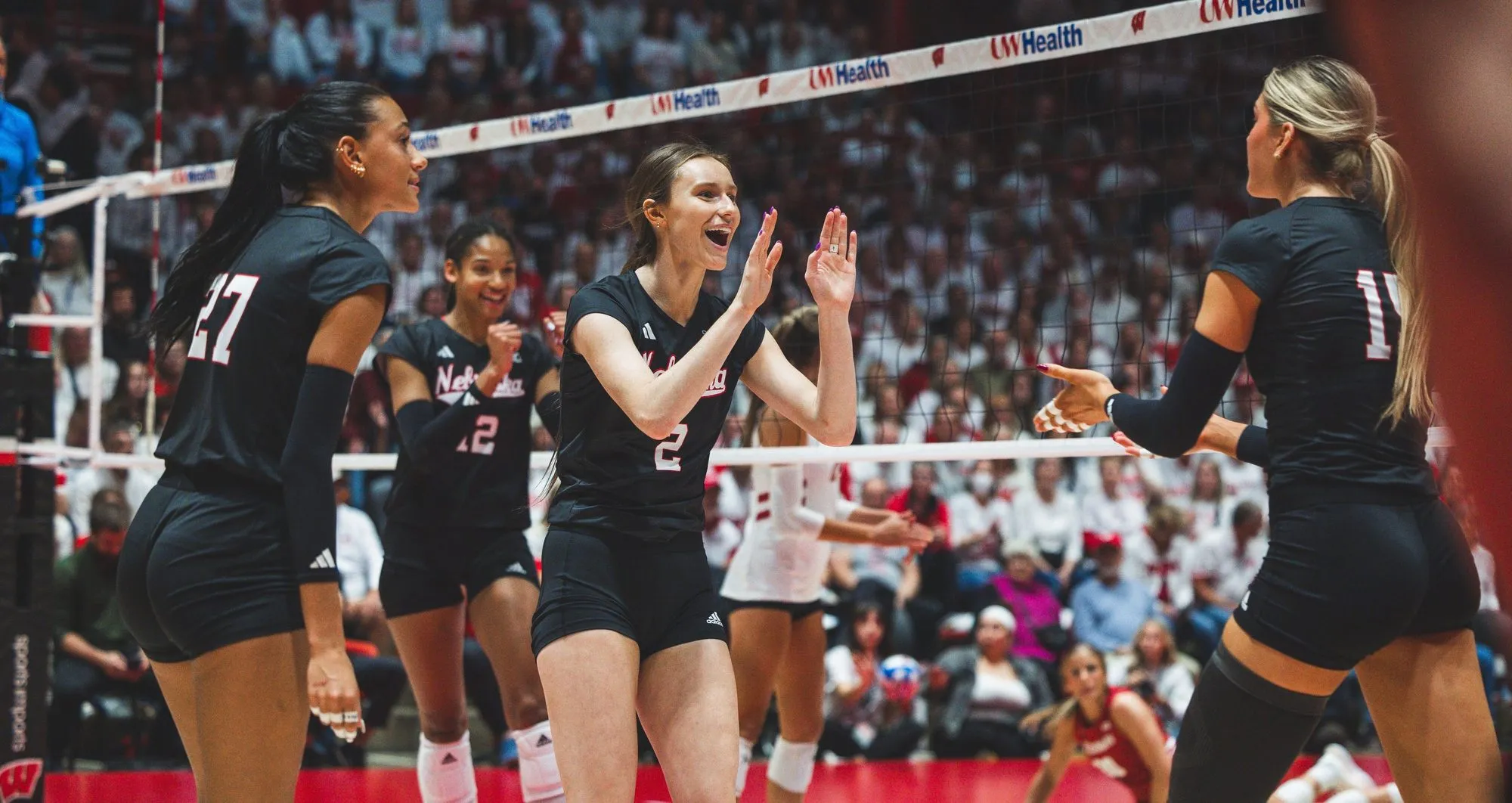Four Nebraska volleyball players in black uniforms celebrating together at the net, hands raised, in front of a packed arena.