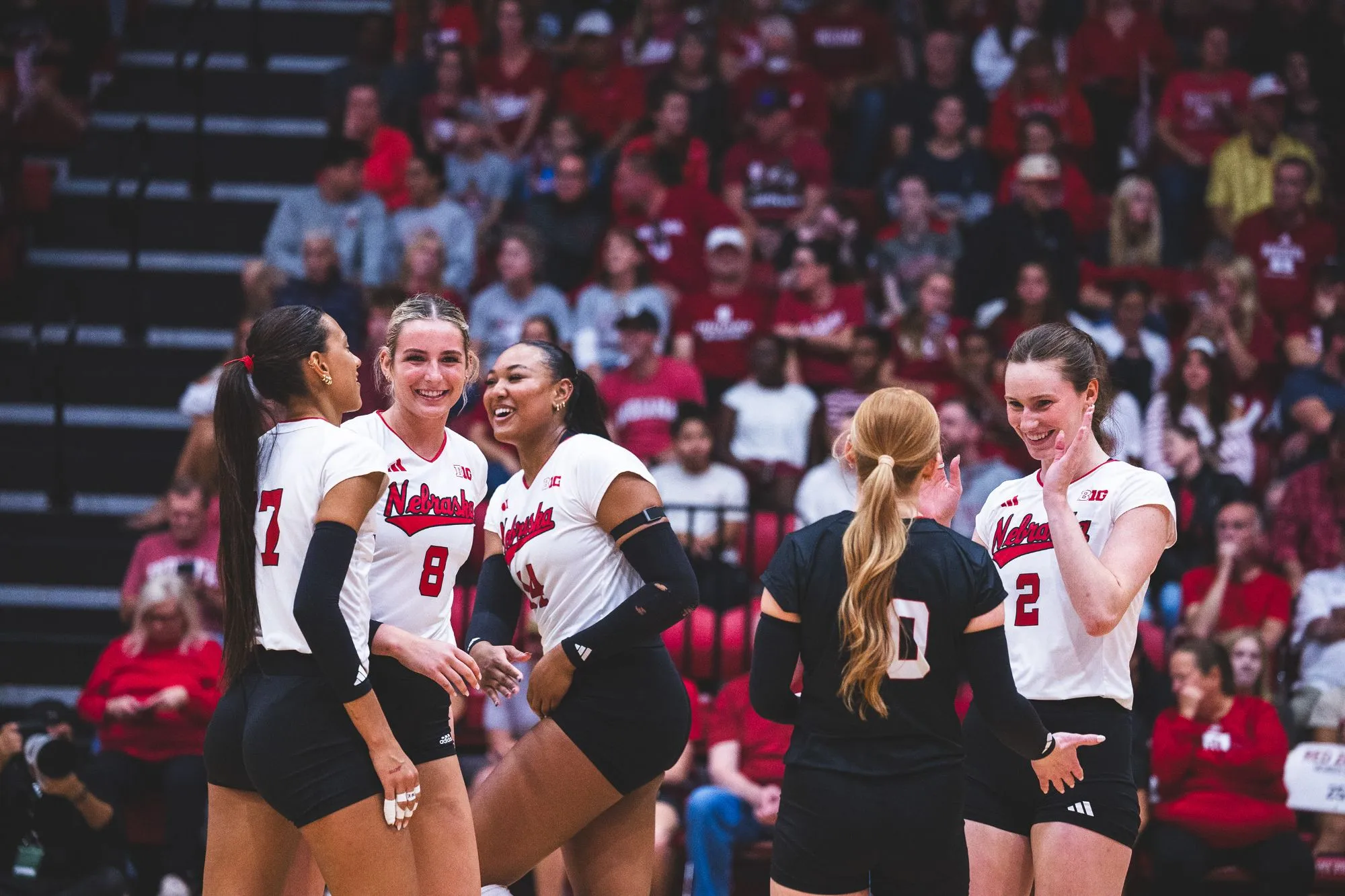 Five Nebraska players smile and gather on the court during the sweep at Indiana's Wilkinson Hall, with a crowd dressed in Indiana red behind them.