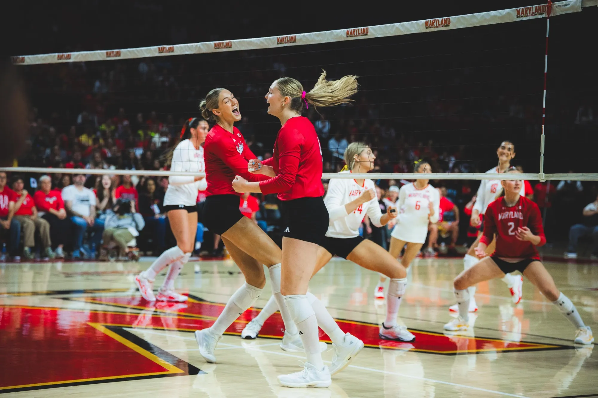 Two Nebraska players in red celebrate at the net during the sweep at Maryland's XFINITY Center, with Maryland players in white and a packed crowd behind them.