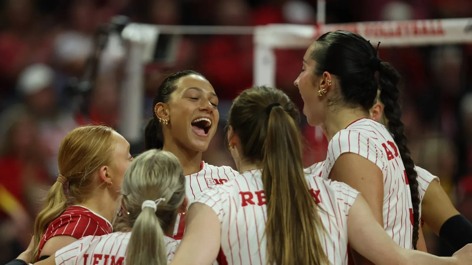 Nebraska volleyball players celebrate a point in a huddle at the net, with Bergen Reilly and Keri Leimbach turned away from the camera.