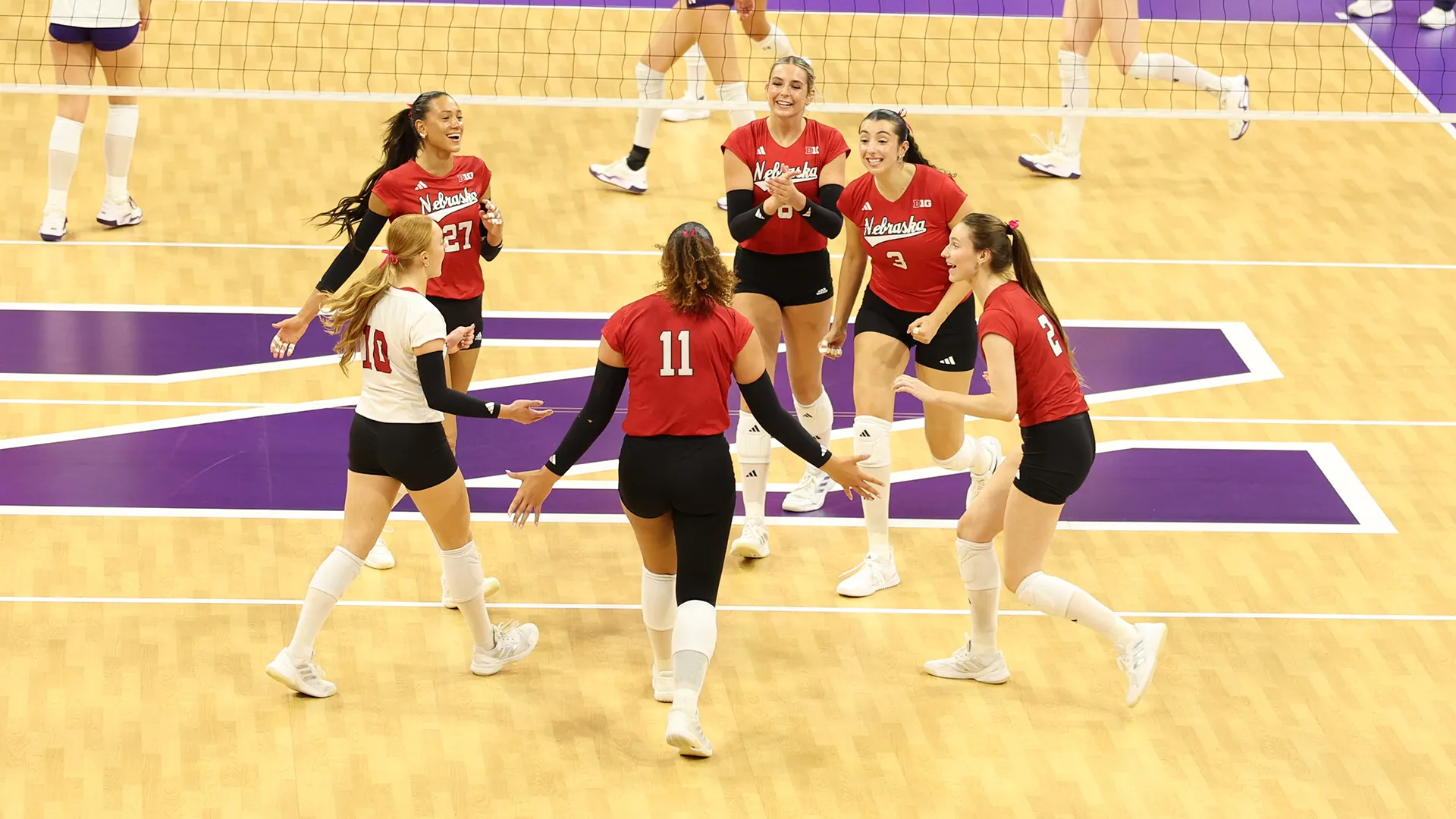 Six Nebraska players, five in red and one in white, smile and step toward each other on the court at Welsh-Ryan Arena, with a large purple N on the wood floor and the net behind them.