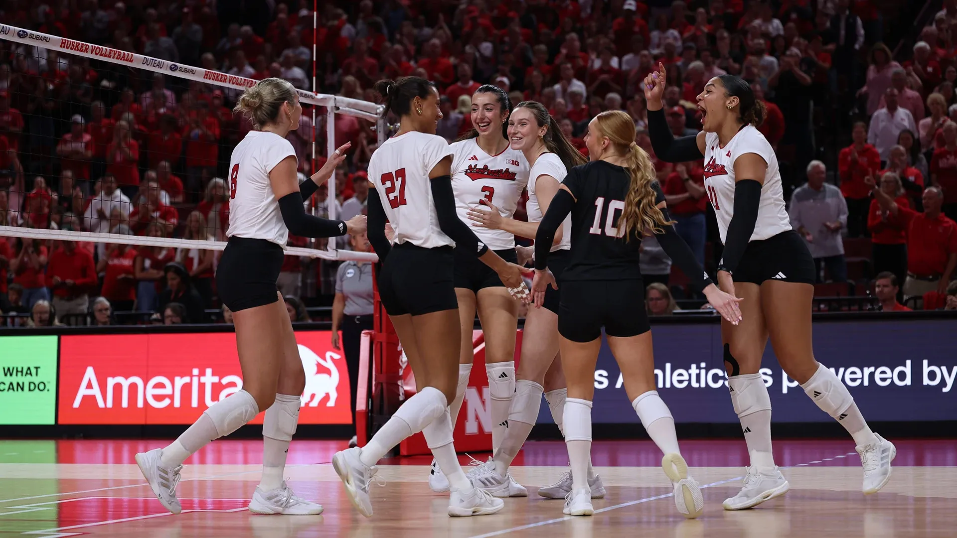 Six Nebraska players gather at the net in celebration, smiling and reaching to slap hands, in front of a packed, red-clad crowd.