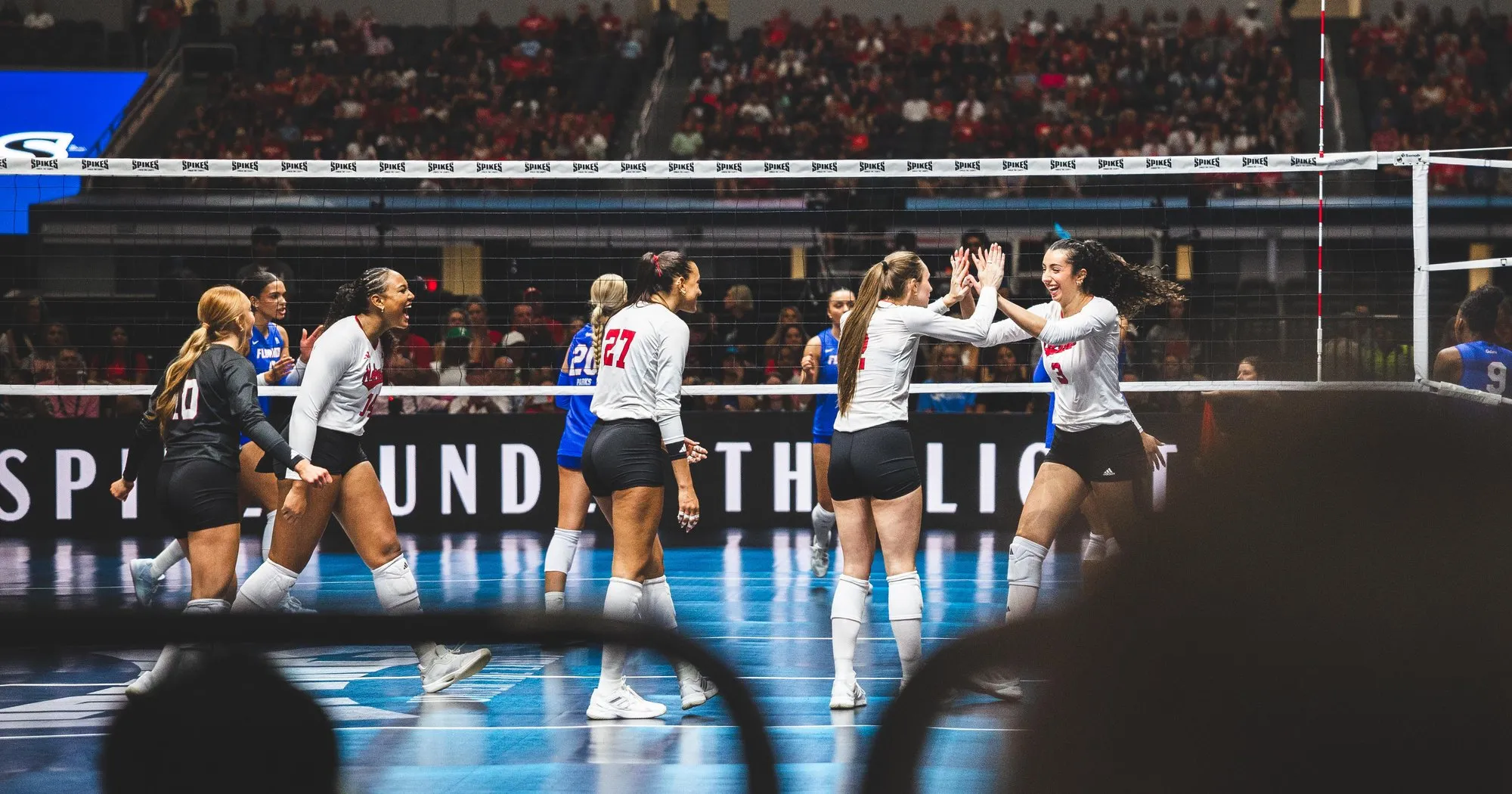 Six Nebraska volleyball players in white celebrating along the net, two of them high-fiving, in front of a packed red-clad crowd with Florida players in blue on the far side.