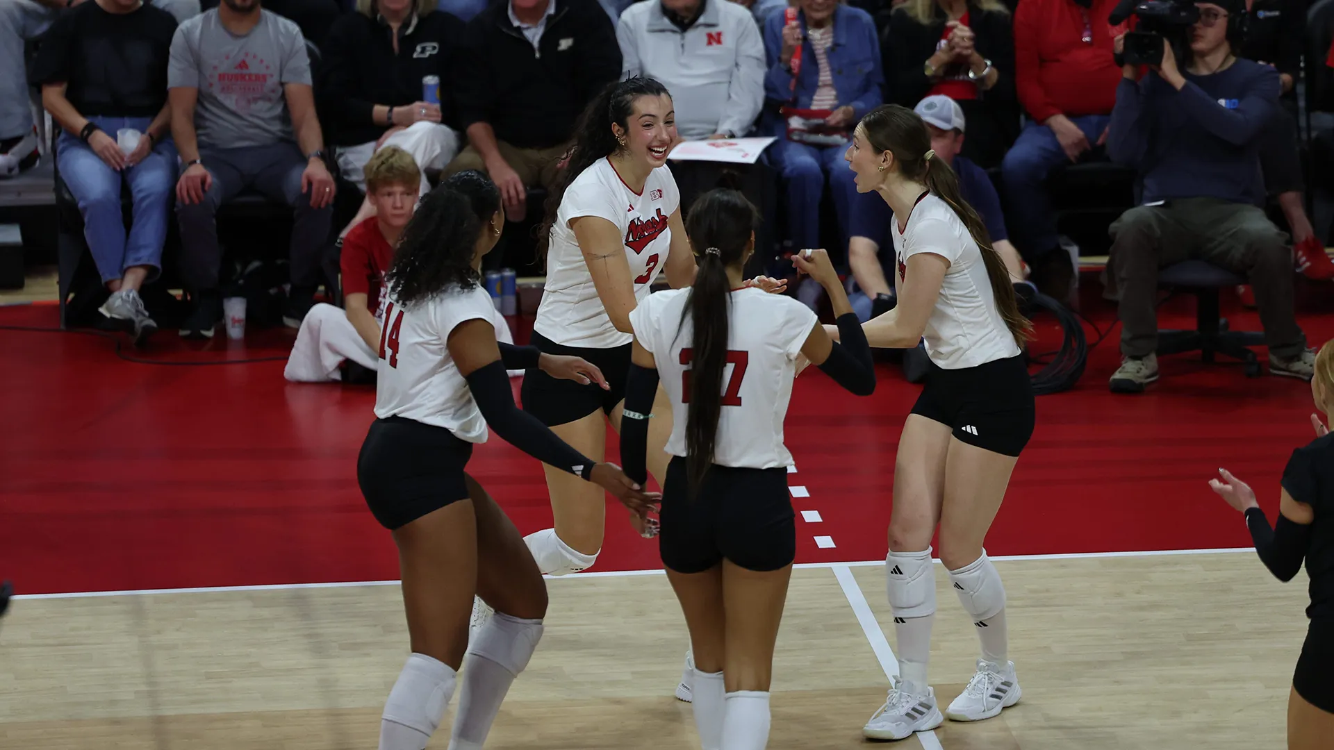 Four Nebraska players in white jerseys, including No. 14, No. 3 and No. 27, smile and come together to celebrate a point on the court at John Cook Arena, with fans in the stands behind them.