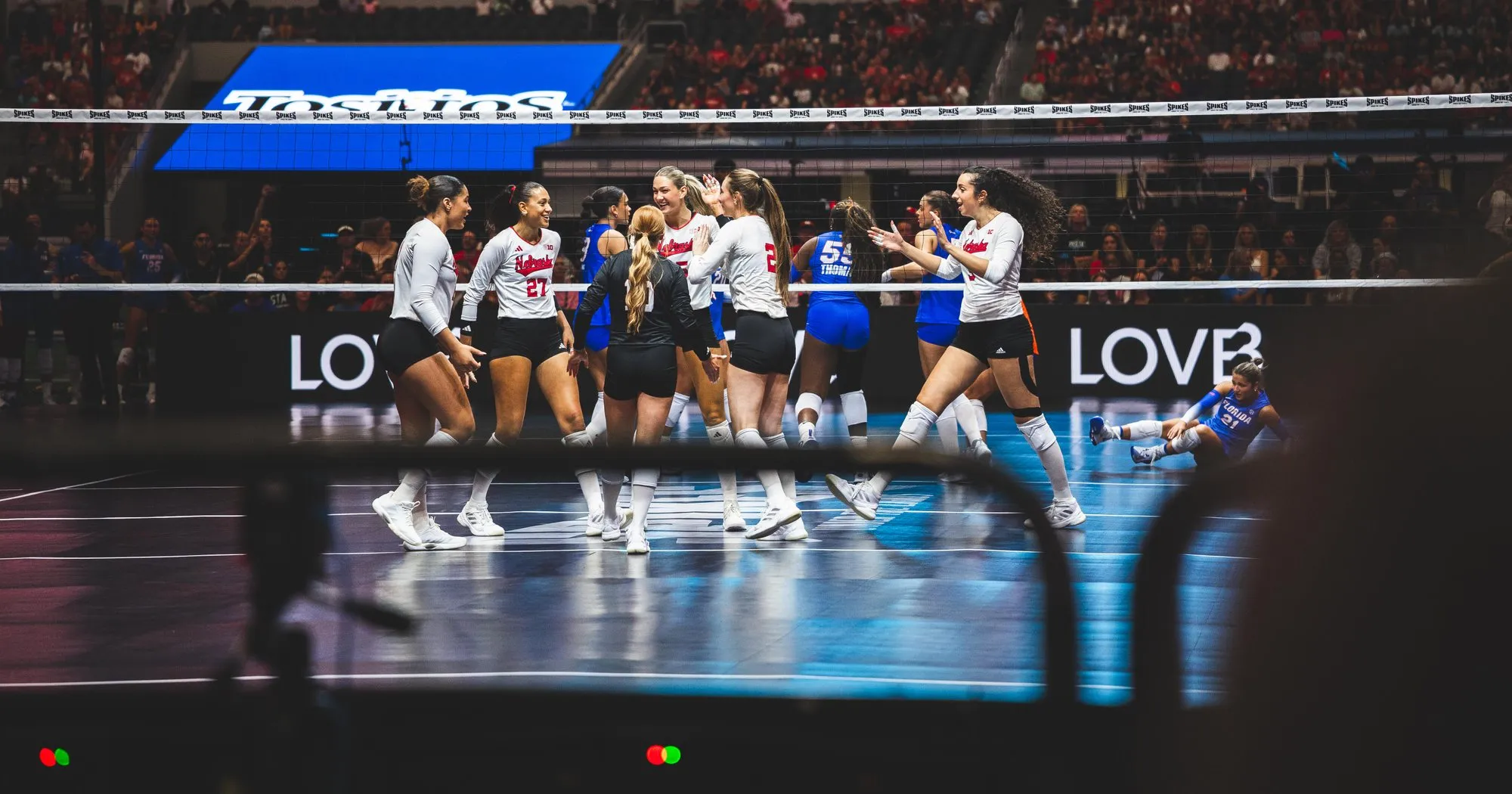 Nebraska players in white gathered at the net celebrating a point, while a Florida player in blue sits on the court behind them, in a packed AT&T Stadium.