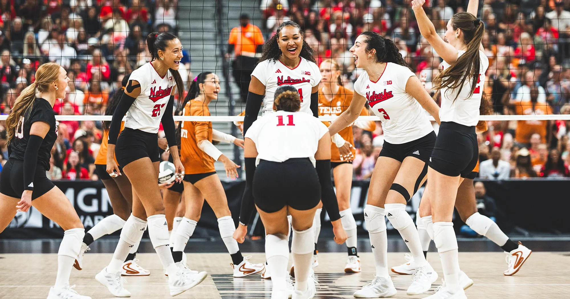 Six Nebraska volleyball players in white celebrating together at the net, with Texas players in burnt orange standing behind them, at T-Mobile Arena.