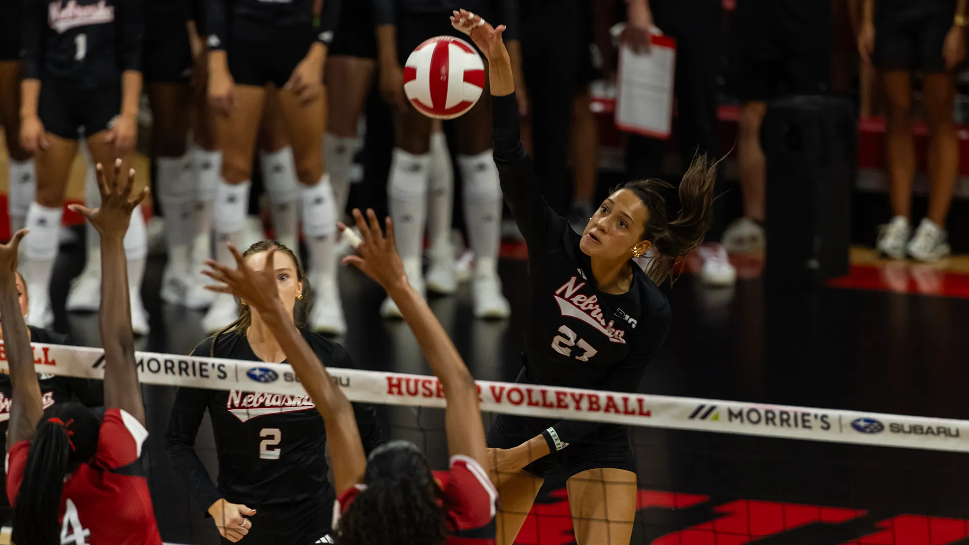 A Nebraska outside hitter in a black jersey leaps and swings at the ball above the net as two Wisconsin blockers in red reach up, with a teammate watching from the other side of the net.